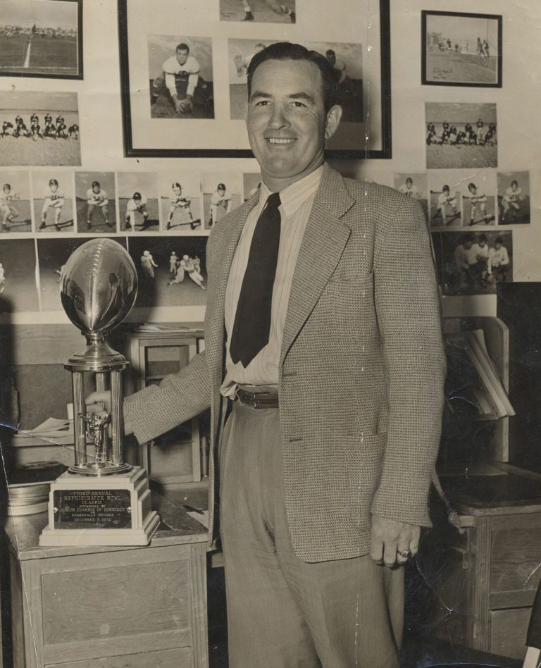 Man in a tweed jacket smiling beside a Refrigerator Bowl Classic football trophy, from the Pete Ragus collection