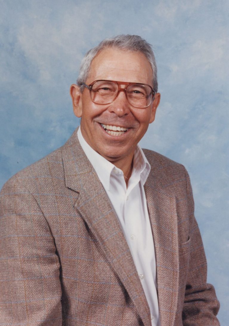 Pete Ragus smiling in glasses, a tweed sport coat and a white shirt in a studio portrait