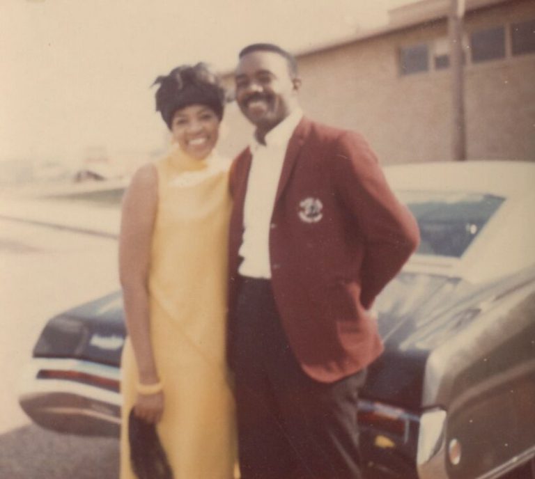 Man in a maroon blazer with a smiling woman in a yellow dress beside a car, from the Prenis Williams collection