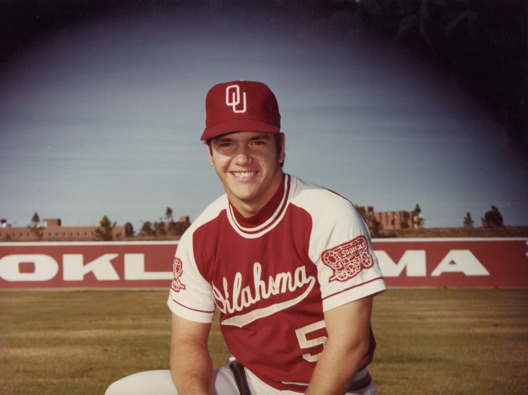 Steve Coleman smiling in an Oklahoma baseball cap and crimson jersey in front of an OKLAHOMA outfield wall