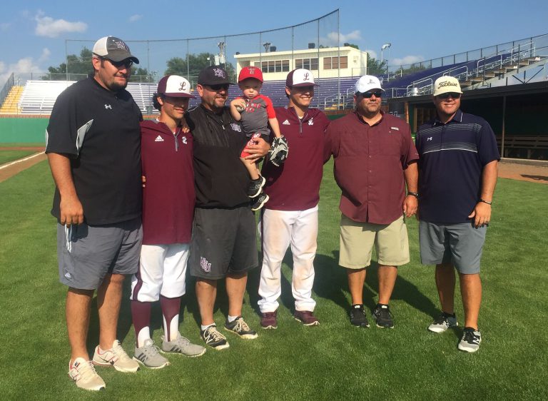 Steve Coleman with a group of coaches, two players in maroon and a toddler posing on a baseball field