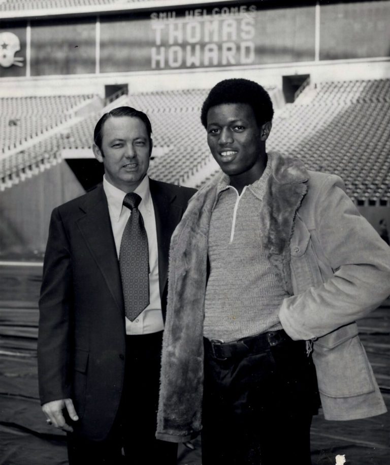 Thomas Howard in a fur-lined jacket beside a man in a suit under an SMU Welcomes Thomas Howard stadium sign
