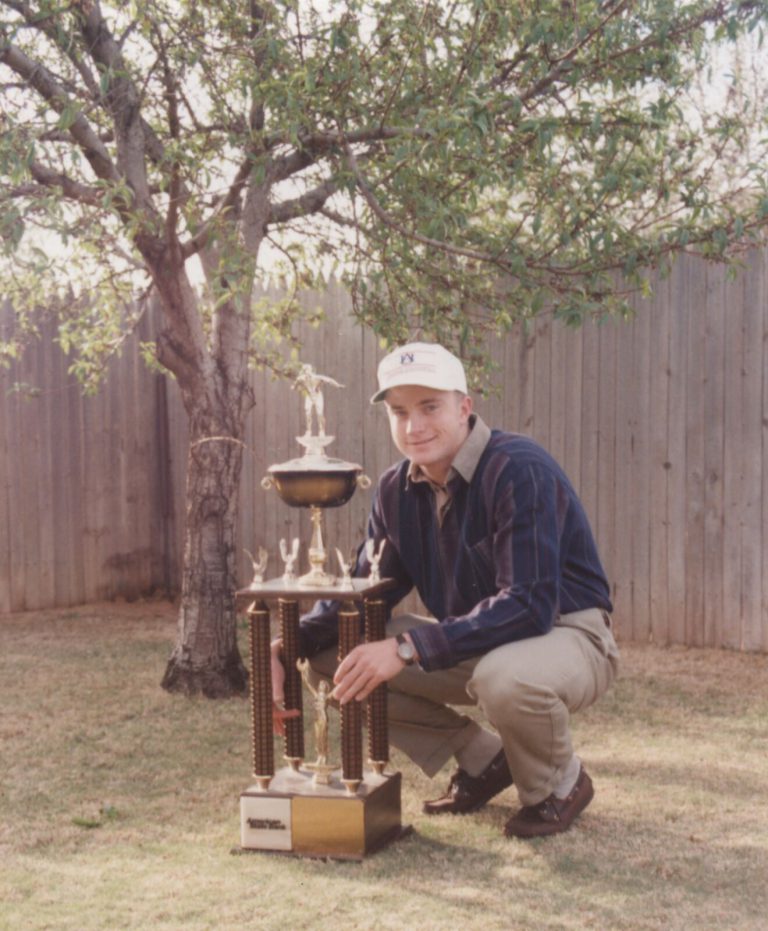 Thomas Klement in an Auburn cap crouching beside a tall trophy under a backyard tree
