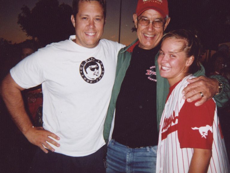 Amanda Parsons, right, in a pinstriped red-and-white jersey with a mustang logo, posing with two smiling men