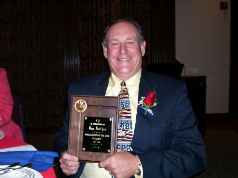Ron Holihan in a navy blazer with a red boutonniere holding an engraved award plaque