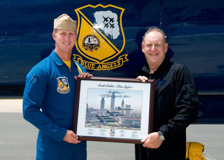 Ron Holihan, right, in a dark jacket holding a framed Blue Angels print beside a Blue Angels pilot after a VIP flight