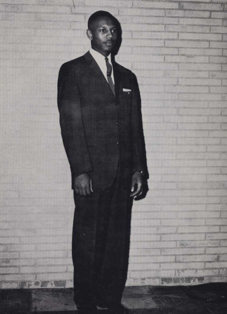 Joe McWilliams stands in a suit and tie in front of a brick wall in a vintage Dunbar High School photo.