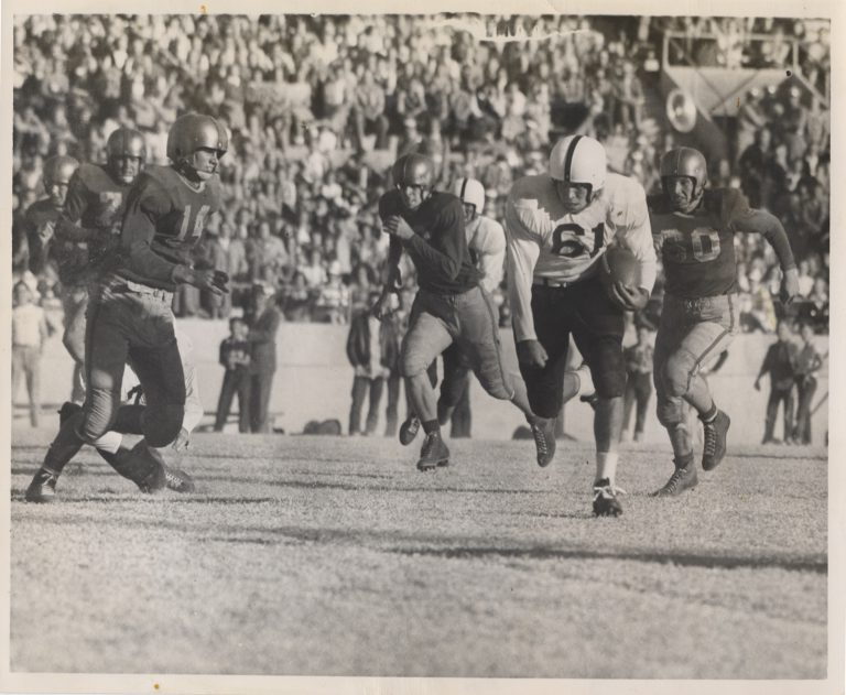 Mac Taylor, number 61, carries the ball past defenders in front of a packed stadium crowd around 1950