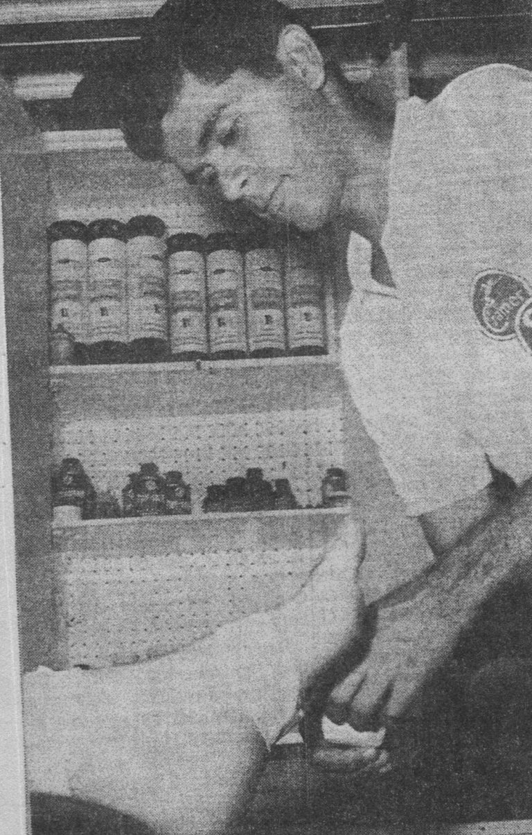 Robert “Prof” Ramos as a student trainer working on an athlete's ankle in front of a shelf of supplies, from a clipping