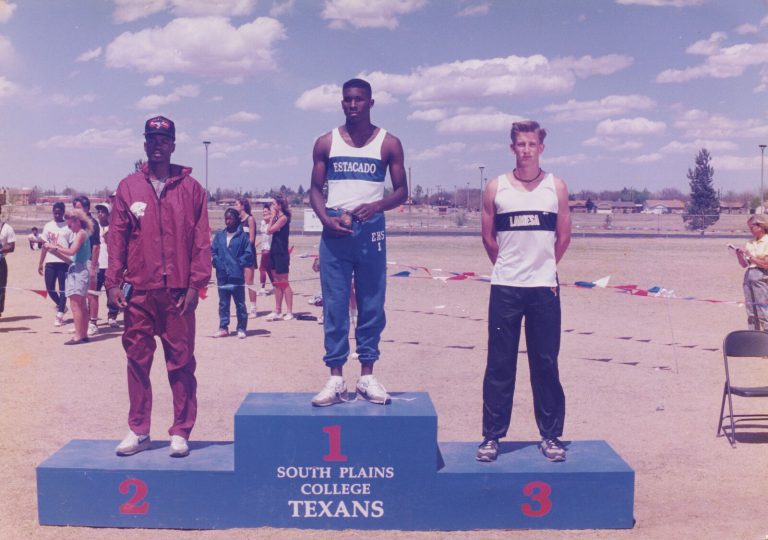 Taje Allen on the first-place step of a South Plains College Texans podium in an Estacado singlet, with two other medalists