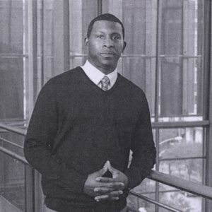 Taje Allen in a sweater and tie, standing indoors in a recent professional portrait.