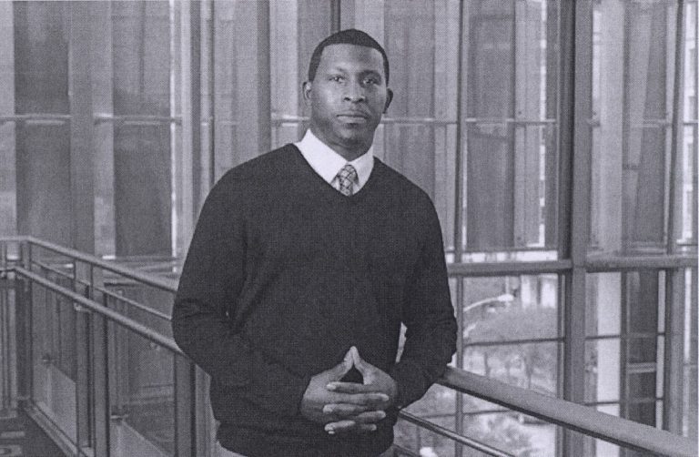 Taje Allen in a sweater and tie, standing indoors in a recent professional portrait.