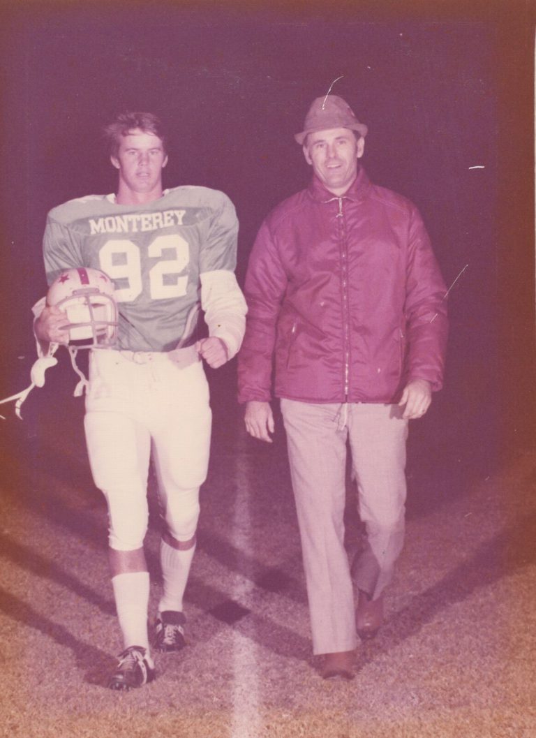 Tim Leslie in a Monterey number 92 football jersey, carrying a helmet and walking the field at night with a man in a jacket